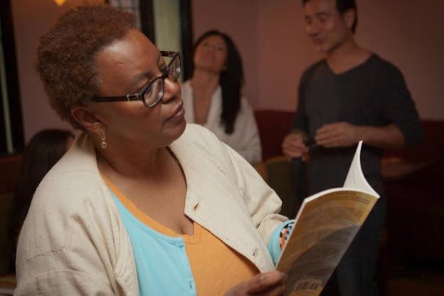 Photograph of a person in glasses and a beige cardigan looking down at an open book they are holding.