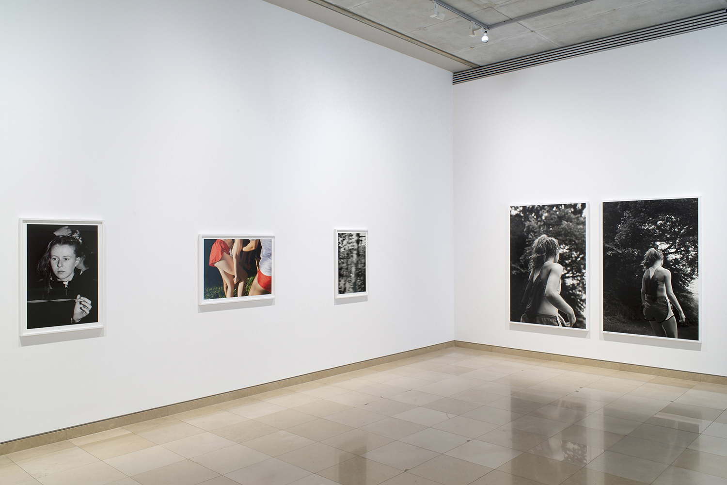 A photograph of two black and white framed photographs in white frames hung next to each other. Surrounded by wide white mat within the frame, the photographs are closeup shots of a subject's face. In both, the subject gazes downwards, shielding their eyes from the sun.