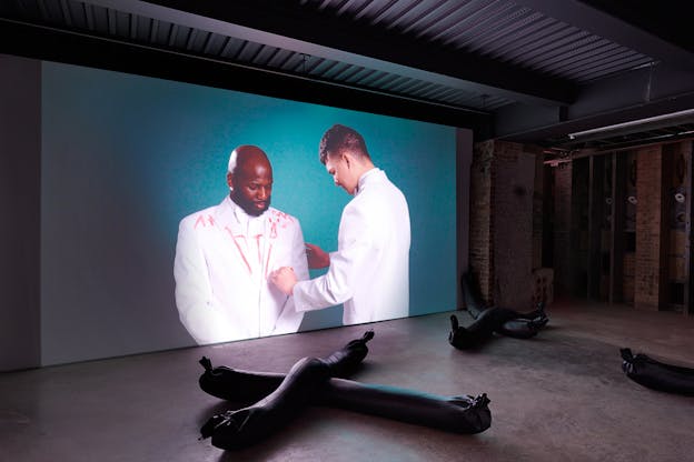 Three soft sculptures comprised of black cylindrical shapes tied off at the end lie in X shapes on a concrete floor. Behind them two performers in white tuxedos are projected onto the wall. One of them is making red marks on the other’s tuxedo.