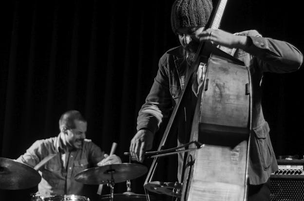 Black and white photograph of two figures playing musical instruments. The figure in the front closest to the viewer fashions a beanie and a beard as they play a bass that is turned sideways to the photo, while the figure in the background on the left grimaces as they play the drums.