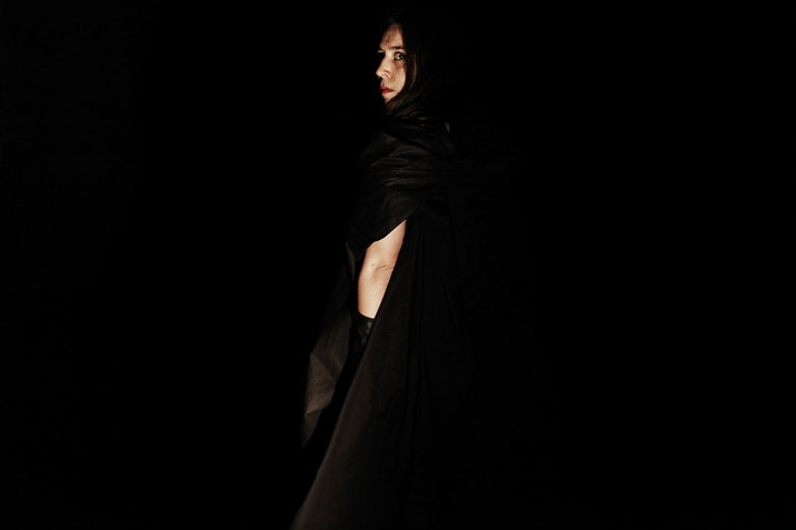 Achugar faces away and turns her head to look forward, cloaked in black clothing that blends into pitch black space.