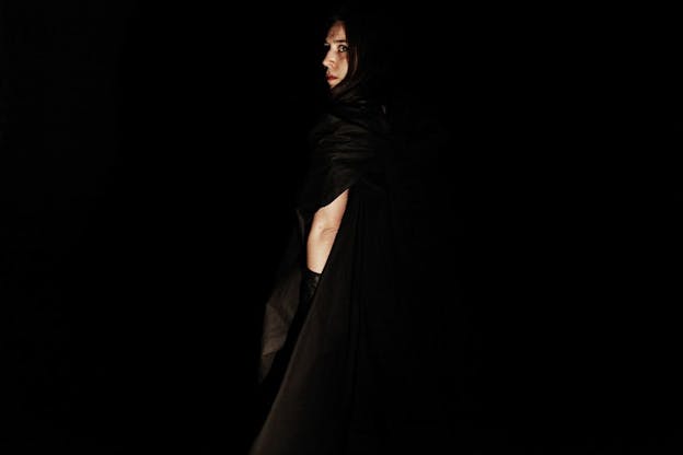 Achugar faces away and turns her head to look forward, cloaked in black clothing that blends into pitch black space.