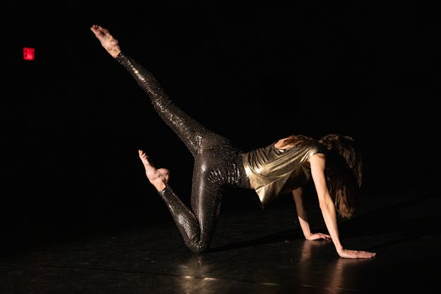 Joanna Kotze performs alone on a dark stage. She is positioned in profile to the camera, head pointed toward the right, face looking down at the floor. She balances as a tripod on both hands and her right knee, with her head, shoulder, and hips in line with one another. As she balances with her knee on the floor, her right foot is raised in line with her hips and her toes are pointed. Her left leg is extended and raised up behind her, foot pointing upwards. She wears dark metallic leggings and a loose gold metallic tunic, her hair falls, covering her face from view. 