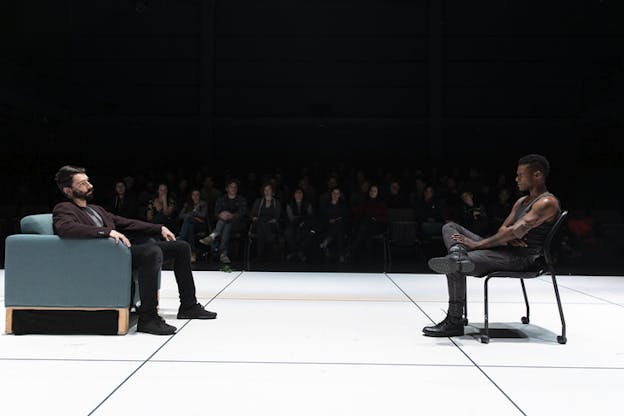 Two performers sit facing each other in a couch and in a chair on a stage with big white tiles. Behind them en face to the viewer, an audience sits in the dark.