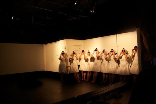 A group of performers in matching white smocks and head scarves adjust their sunglasses, standing against a yellow-lit stage wall. 