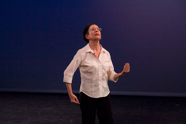 Performer dressed in a white button-up, a black cap, and glasses looks upwards and curves their fingers forward and to their side on a stage with a black floor and a deep purple backdrop.