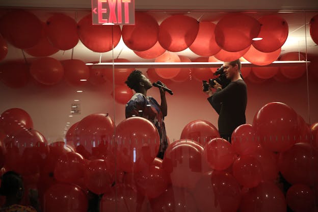 Autumn Knight performs in a glass enclosed space filled with red ballons which cover the floor up to her waist and the ceiling. She wears a black shirt with a red and white pattern and holds two microphones, speaking into them and facing a person holding a video camera opposite her.  
