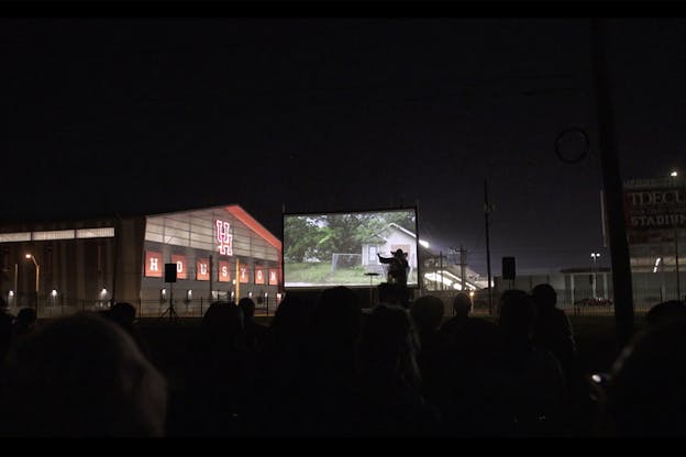 A crowd watches a performer on a dimlight scene, a projection of a house surrounded by green behind the performer. Behind the scene on the left stands a building with the word 