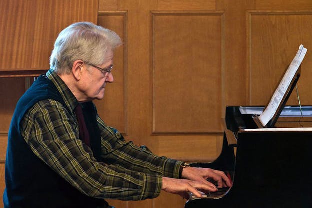 Gordon Mumma plays the piano in a wooden space. He has grey hair and wears a plaid shirt, dark vest, and glasses. He has a serene expression on his face. 