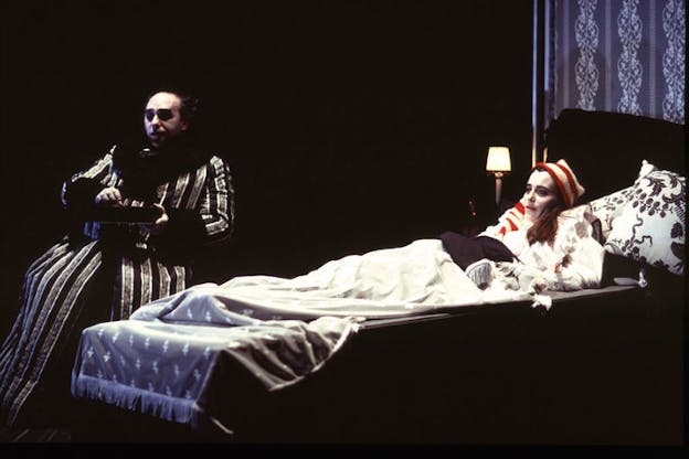 A woman with a sickly pale face lays in bed while a man clad in dark clothing, white makeup and dark eyeshadow sits besides her looking in the distance.