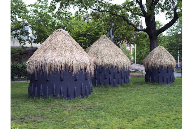 Load-shaped round house structures with black walls and a straw triangle roof in a park with green grass and trees.