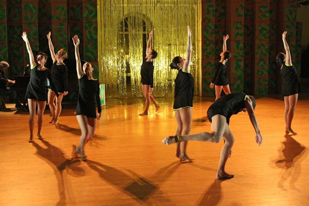 On a dimly lit stage with a honey-colored wooden floor and curtain backdrop of gold tinsel, performers dressed in black move around the stage in a circular form raising and looking up at one arm.