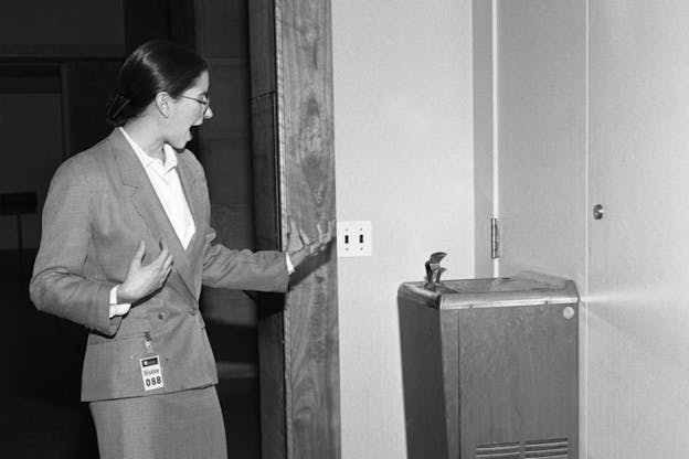 A black and white picture of a figure dressed in a suit and pencil skirt, facing a water fountain with one hand clawed upwards in front of them. 