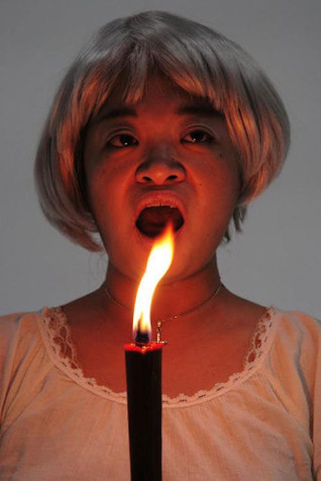 Nexus6 performs, wearing a white lace shirt, short blonde wig, lavender eyeshadow, and a necklace of a cross. In front of her, there is a thin lit candle. She opens her mouth, as if mid-speech. 