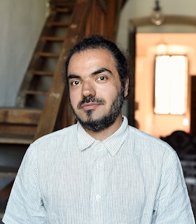 A photograph of Shahpour Pouyan from the ribs up, looking at the camera. He has dark hair, a moustache, and a short beard, and wears a button-down shirt with thin stripes.