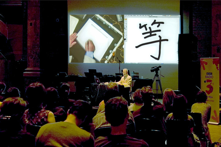 Yasunao Tone stands behind a black desk in front of an audience. Behind Yasunao Tone, there is split a projection of his hands drawing something on the left and an alphabetical character on the right. The stage and audience is bathed in low reddish light.