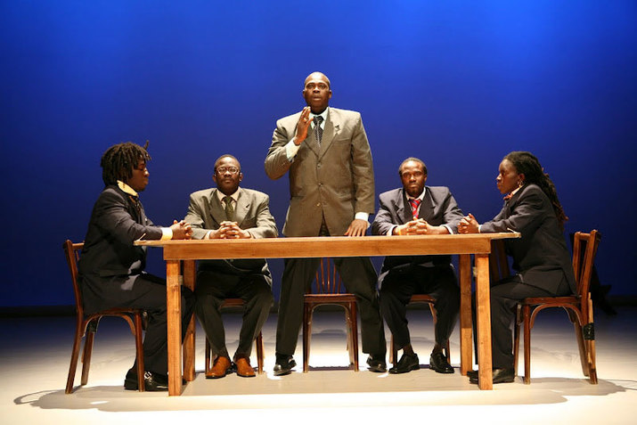 Five performers wearing grey and brown toned tuxedo outfits are gathered around a wooden table. The one in the middle stands up and raises their right hand while the rest of them are seated with their interlocked fingers resting on the table. The stage has a beige color floor and a blue gradient backdrop.