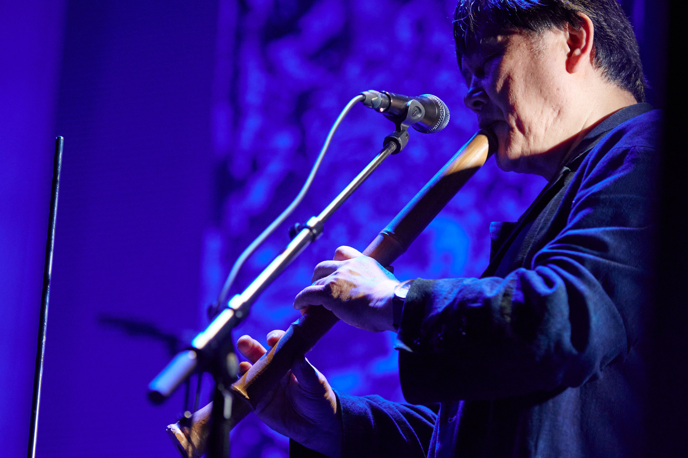 Koichi Makigami performs on a stage awash in blue light. He stands in profile on the right side of the frame facing the left. He stands before a microphone and plays a large wooden flute. He is wearing a dark long sleeve shirt. 