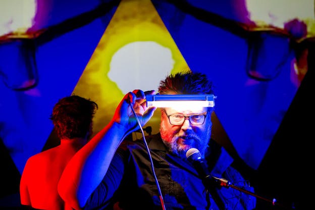 Performer in front of a microphone holds a white LED light above their spectacled eyes. Their body and the back of a performer standing behind them are bathed in neon red and blue light which blends into a screen behind them projecting two abstract spectacle rims on each side of a molten yellow pyramid. 