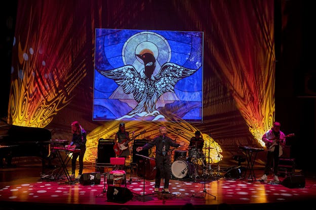 Performers play instruments and sing on a red lighted stage. Behind them a yellow and orange background centers on a blue square illustrating a bird with its wings open.