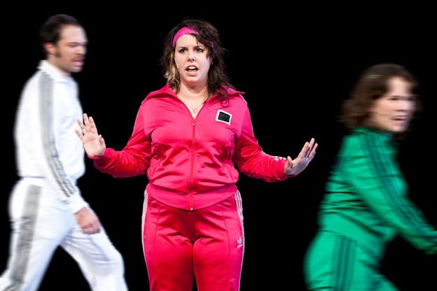 An in-focus performer dressed in a hot pink Adidas tracksuit and headband faces forward saying something with their hands extended and elbows bent while on both of their sides, two blurred figures move. 