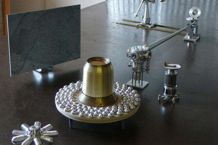 A bell shaped golden object rests on the center of a circular wooden plate. Around the gold object, there are numerous silver spheres. Besides the wooden plate, there is a blue toned map annotated with red. Around the wooden plate, there are different parts of silver machinery.