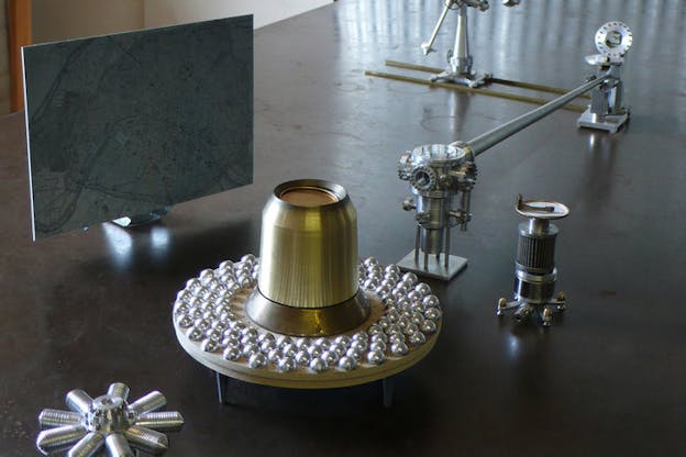 A bell shaped golden object rests on the center of a circular wooden plate. Around the gold object, there are numerous silver spheres. Besides the wooden plate, there is a blue toned map annotated with red. Around the wooden plate, there are different parts of silver machinery.