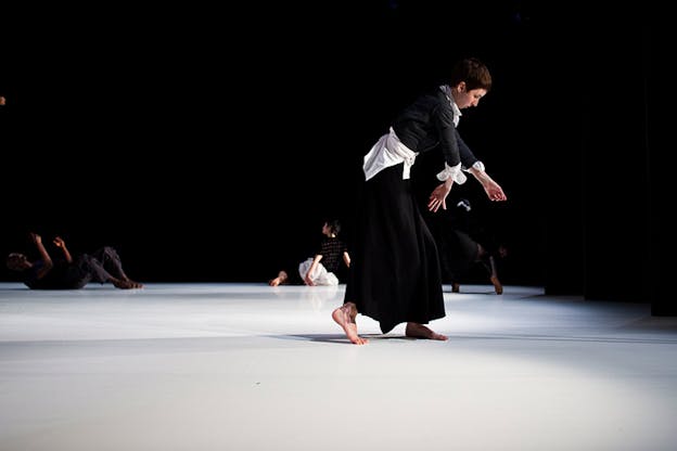 On a black-lit stage with an illuminated white floor, a performer dressed in long black and white garb bends slightly forward with one back foot in demi-pointe, their arms extended and slightly bent. In the distance, performers dance on the floor. 