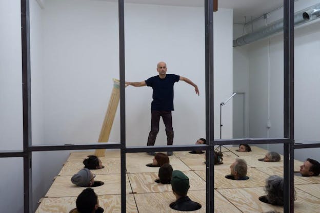 Performer popping and holding a tilted pedastal standing on a wooden floor cut into circles through which people are sticking up their heads. 