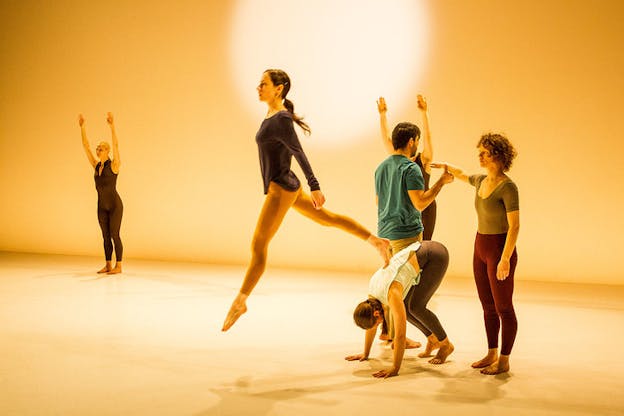On a yellow-lit stage one performer leaps off the back of a person bending in front of three performers striking poses in a cluster, in the distance a performer in a black unitard raises their arms straight above their head.