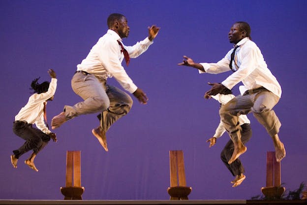 Four performers each wearing a tucked white shirt, a patterned necktie, and a pair of fitted pants leap into the air facing each other. Three wooden chairs  with tall backs are placed against a purple gradient backdrop.