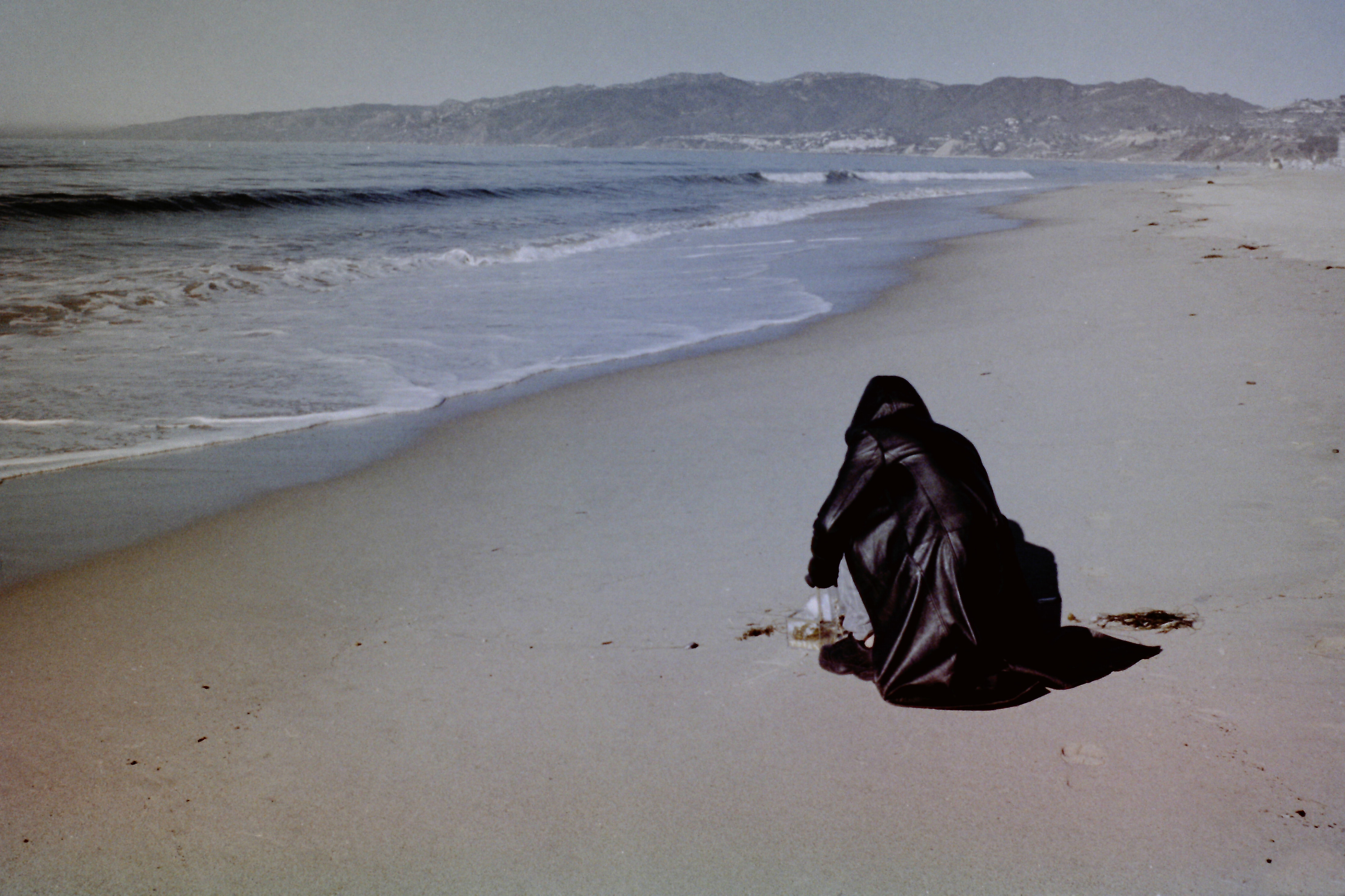 Angel Lartigue crouches on the sand in front of a clear acrylic box with her back facing the camera. She wears a hooded black leather jacket. Behind her, the shoreline extends into the distance where it eventually meets a mountain range as waves crash against the beach.