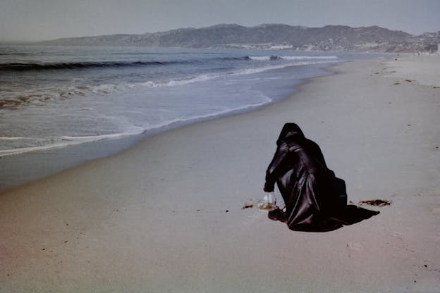 Angel Lartigue crouches on the sand in front of a clear acrylic box with her back facing the camera. She wears a hooded black leather jacket. Behind her, the shoreline extends into the distance where it eventually meets a mountain range as waves crash against the beach.