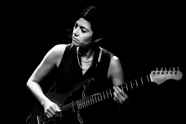 A black and white photo of Ava Mendoza playing a dark electric guitar against a black background. Visible from the hips up, their body is facing the camera, head turned towards the left and eyes gazing downward. Their right arm is bent as their hand strums the strings of the guitar, and their left hand holds the strings on the guitar neck. They are wearing a dark, sleeveless v-neck top and two chain necklaces.