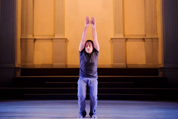 Gries on a blue and yellow lit stage closing his eyes, bending his knees, and raising his arms straight above their head. 