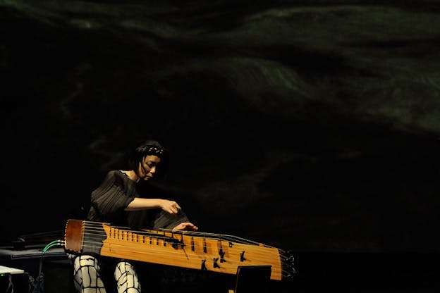 Jin Hi Kim leans over a komungo to play it. She wears a pleated black blouse and black and white patterned pants. The background is all black.