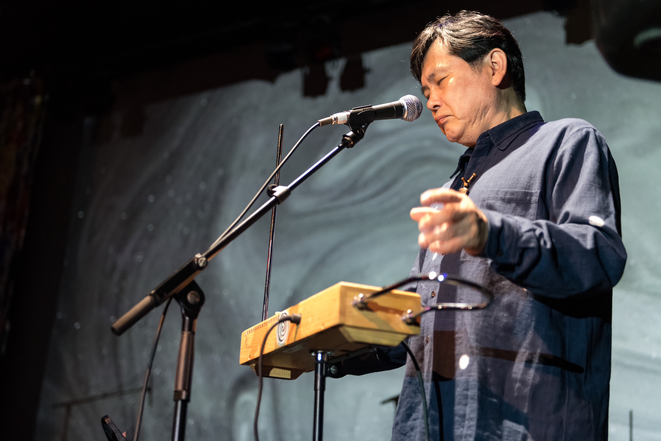 Koichi Makigami performs on a stage in front of a projected abstract silver background. He is standing in the right half of the frame with his body turned towards the left, behind a theremin and a microphone. His eyes are closed and his left arm is bent, with his hand blurred in motion.  