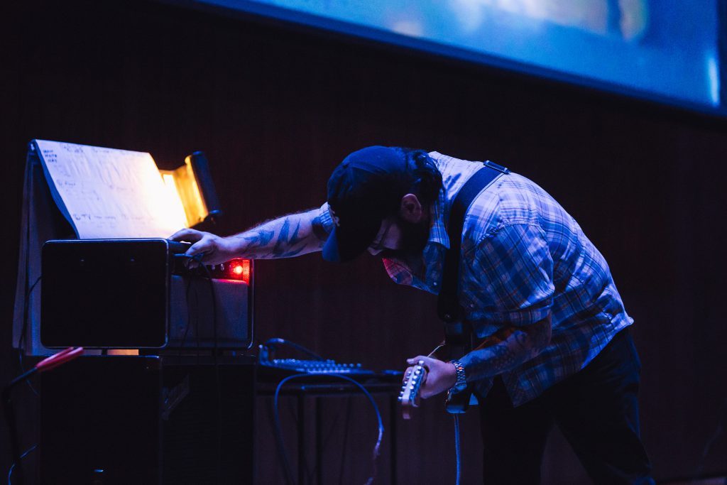 Nathan Young performs awash in blue light, with a soundboard and amplifier by his side, a projection screen visible behind him. He is centered in the image, leaned over to the left to adjust a dial on his amplifier. He wears a guitar strapped over his shoulder and his face is turned towards the ground. He is wearing dark jeans, a shortsleeved blue and white plaid button up, a dark baseball cap, eyeglasses, and a silver watch on his left wrist.