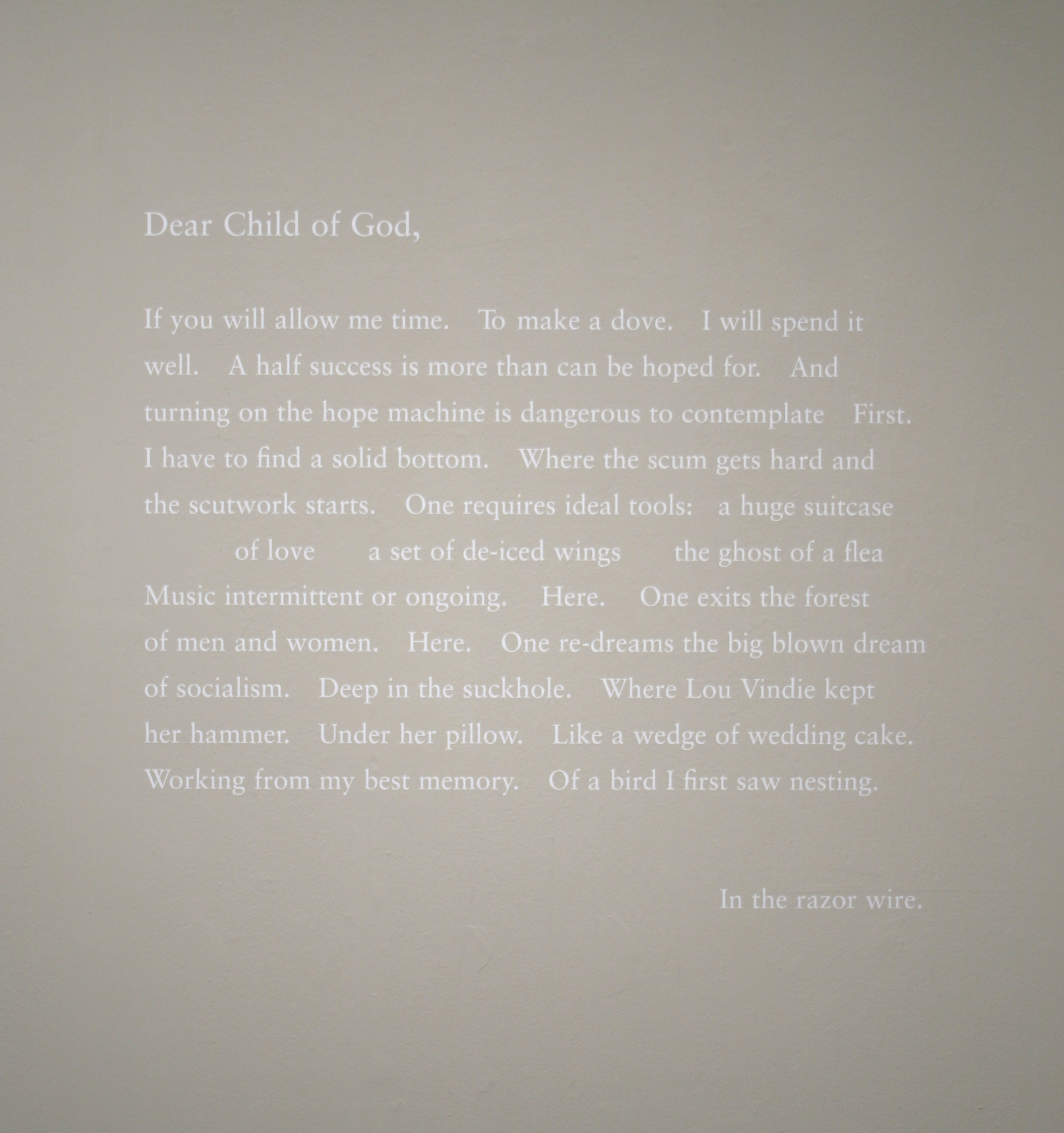 A close up exhibition photograph with a poem written in white text against a white wall. The poem begins with 