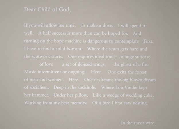 A close up exhibition photograph with a poem written in white text against a white wall. The poem begins with 