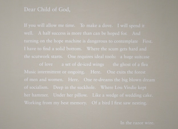 A close up exhibition photograph with a poem written in white text against a white wall. The poem begins with 