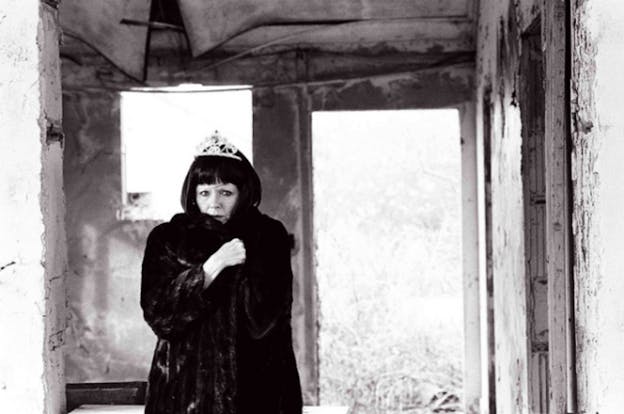 A black and white image of a performer wearing a thick fur coat and a plastic tiara, looking concerned and standing in the middle of a bare room. They wrap their arms across their chest to close the coat and look directly at viewers. The walls behind them are covered in peeling white paint. They are in a small room with openings for a window and an entryway.