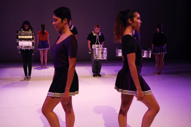 On a purple-lit stage two performers dressed in cheerleader skirts face away from each other, in the background performers hold marching drums and performers dressed in cheerleader skirts face the back wall.