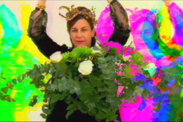 A performer in a mesh black dress and a gold headband raises their arms in front of a bouquet of eucalyptus and white roses. In the background, their image is repeated in various neon hues. 