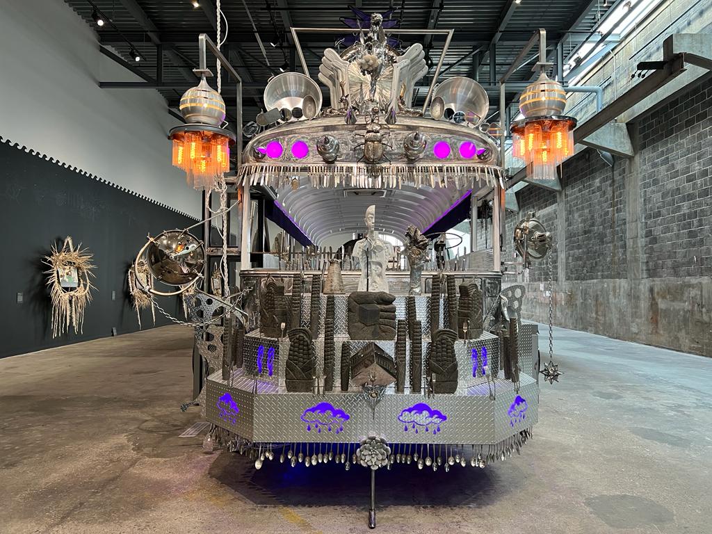 The front view of a large silver sculpture constructed around the skeleton of a schoolbus. The nose of the bus faces the camera and is made from diamond tread metal sheets arranged in three tiers, lined with silver eating utensils. On the tiers are purple illuminated drawings, clay idols in the shape of ears of corn, and other miscellaneous items. Above the windshield are purple lights, spiked accents, and beetle sculpture centered in the middle. The top of the bus is lined with various detached vehicle mirrors, pots, and behind them are a range of larger metal sculptures. Two large hanging lights are fastened to either side of the bus with glowing orange bulbs. 