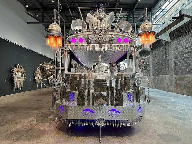 The front view of a large silver sculpture constructed around the skeleton of a schoolbus. The nose of the bus faces the camera and is made from diamond tread metal sheets arranged in three tiers, lined with silver eating utensils. On the tiers are purple illuminated drawings, clay idols in the shape of ears of corn, and other miscellaneous items. Above the windshield are purple lights, spiked accents, and beetle sculpture centered in the middle. The top of the bus is lined with various detached vehicle mirrors, pots, and behind them are a range of larger metal sculptures. Two large hanging lights are fastened to either side of the bus with glowing orange bulbs. 