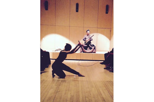 A dancer's shadowed body pictured in profile, blurred in motion, kneeling with one leg and both arms outstretched. On an elevated stage behind, a performer in a robe plays the electric guitar. 