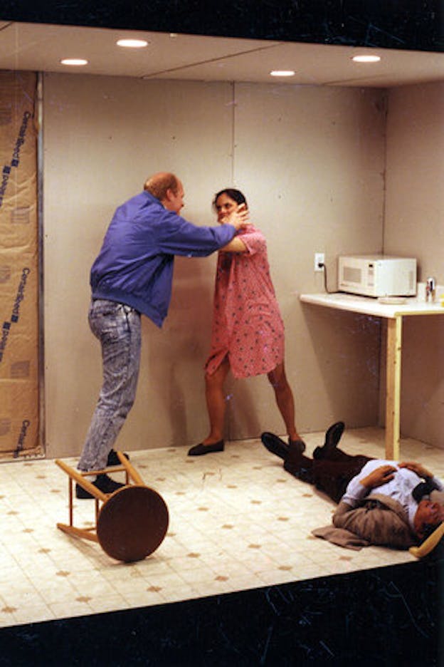 Three performers stand in a grey room. Two performers appear locked in a fight. One of them wears a blue jacket and distressed jeans. The other wears a pink knee length dress and black ballet flats. These performers look intently at one another and hold each other by the neck. A third performer in a grey suit and a cowboy hat lays on the ground immobile.