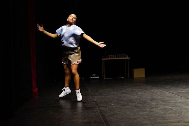 A performer dressed in a blue t-shirt and beige shorts mid-jump with their arms open facing upwards.
