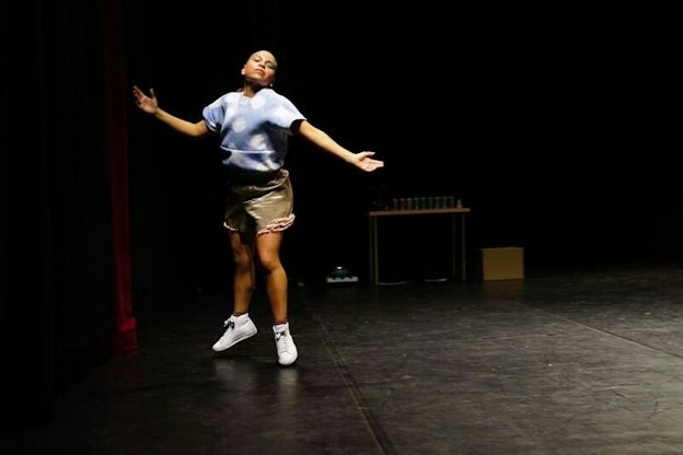A performer dressed in a blue t-shirt and beige shorts mid-jump with their arms open facing upwards.